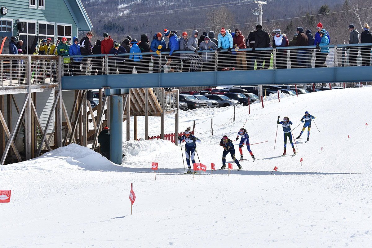 Five cross-country skiers on a race track, with a crowd of people on a bridge overhead.