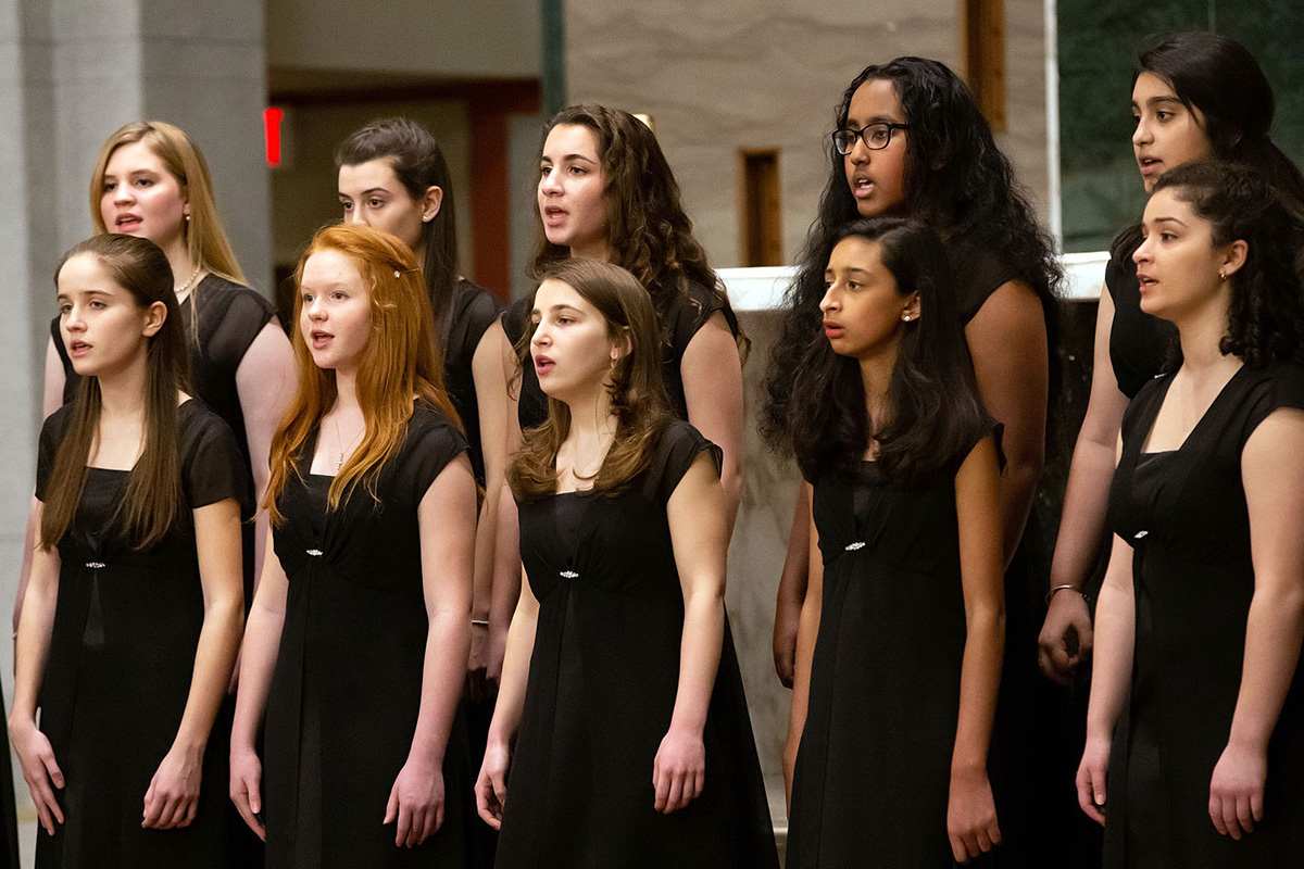 Ten young women, with more out of frame, are dressed in matching black dresses and sing together in unison.