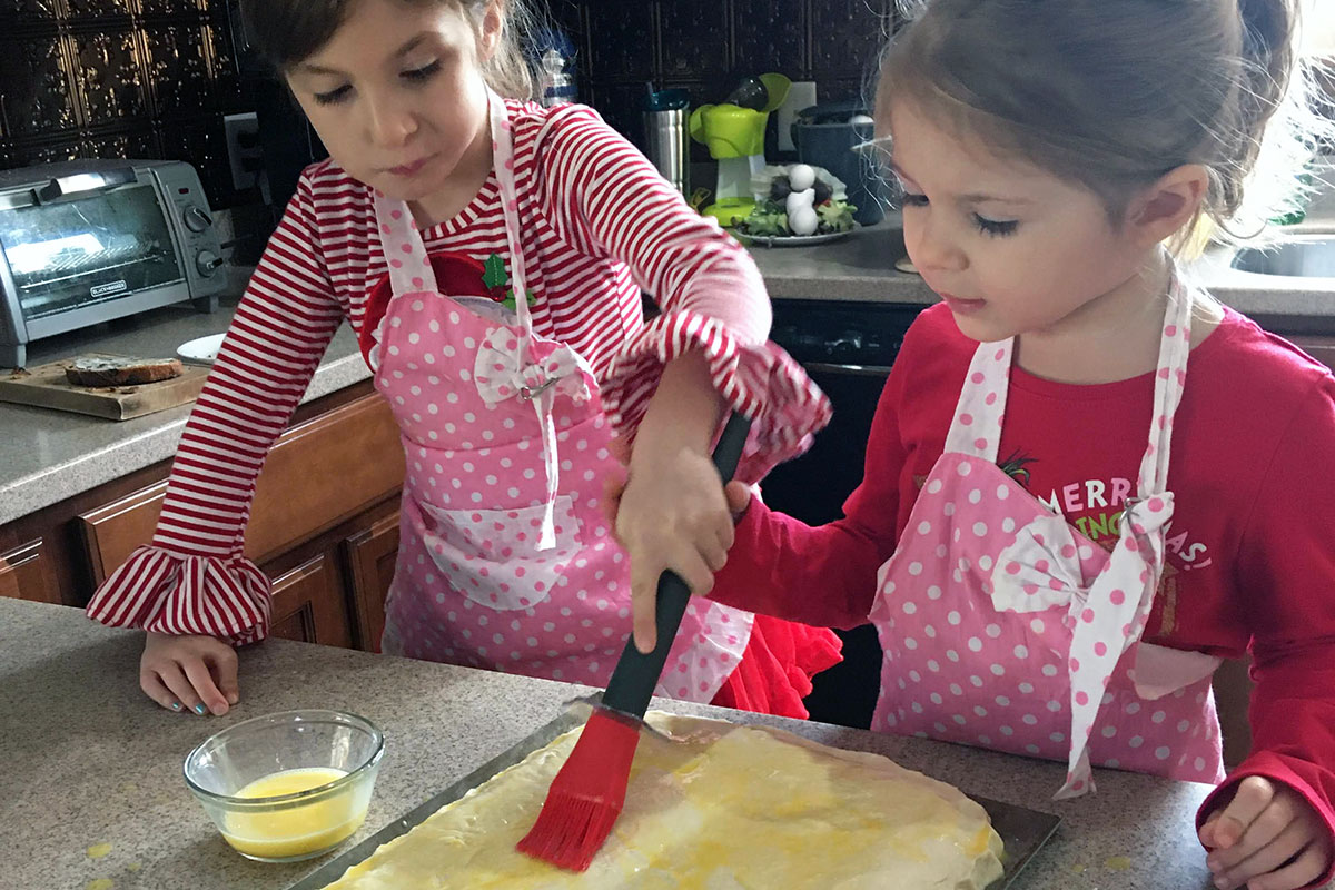 Two young girls in pink polka dot aprons both hold onto the same pastry brush, brushing the top of a slab of dough.