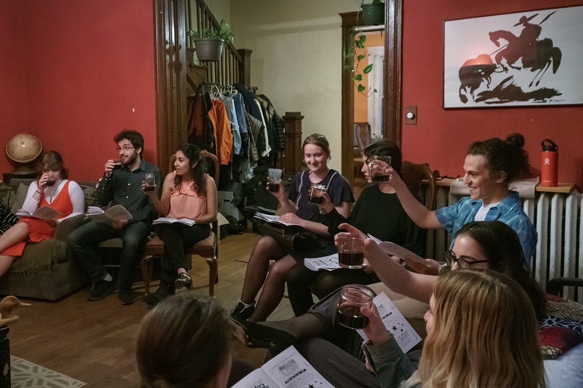 A group of young people raise glasses and jars of red wine, sitting in a living room in mismatched chairs.