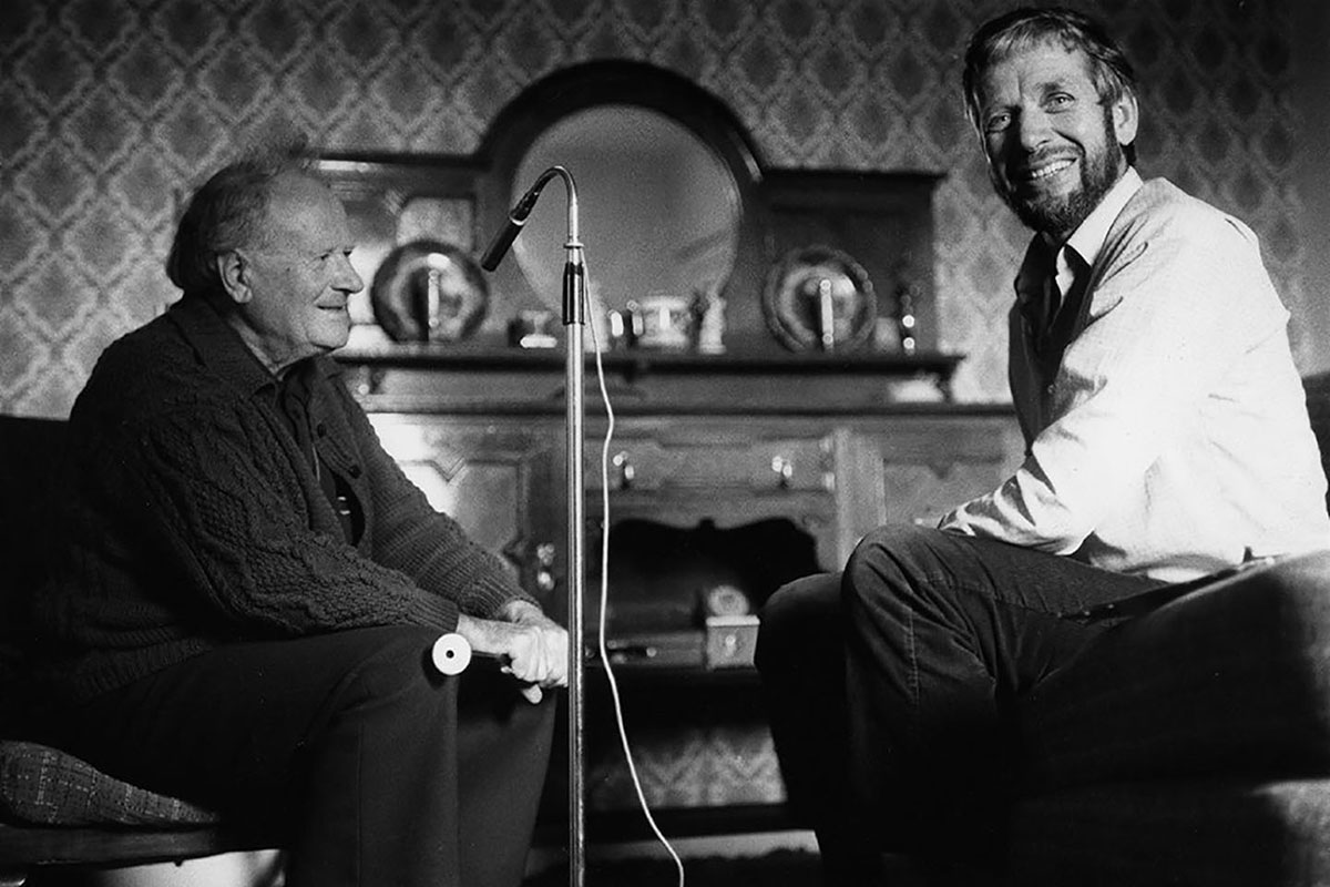 Two men sit across from each other a living room, with a microphone set up between them. The man on the right, dressed in white dress shirt, turns to the camera, smiling. The other, in a dark cardigan, holds a musical instrument, smiling at the man across from him. Black-and-white photo.