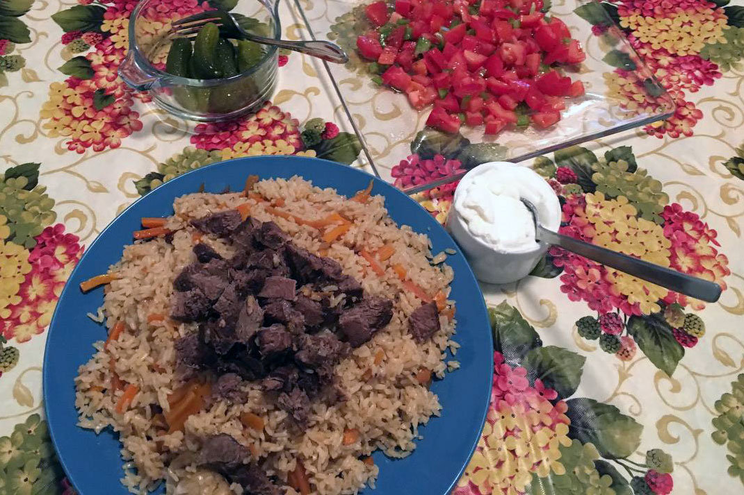 Plate of light brown rice, mixed with orange carrots and topped with chunks of browned meat, alongside a small bowl of yogurt or sour cream, a plate of tomato salad, and a bowl of cucumber pickles.