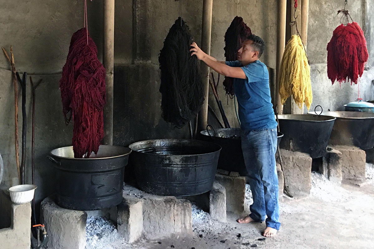 A man in blue jeans and T-shirt, with no shoes, handles a large skein of dark-colored yarn hanging above a pot of dye. Other skeins, in reds and yellow, hang above other pots lined up.