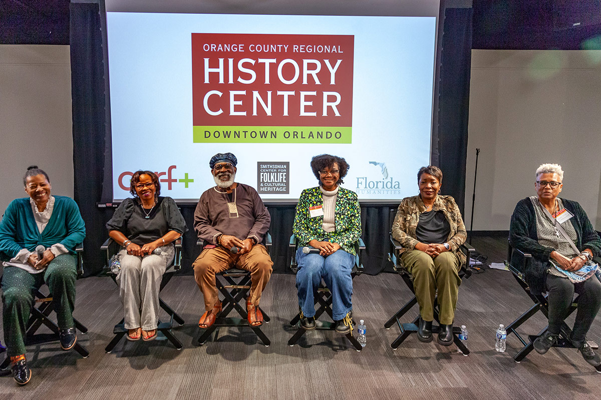 Six African American people sit in chairs lined up in front of a projection screen, all smiling with their hands in their laps. 