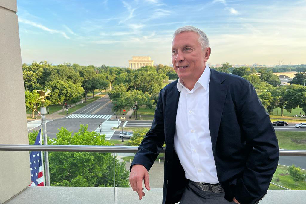 A man with short gray hair, wearing dark blazer and white shirt, smiles as he leans against a balcony railing, with trees and the Lincoln Memorial in the background.
