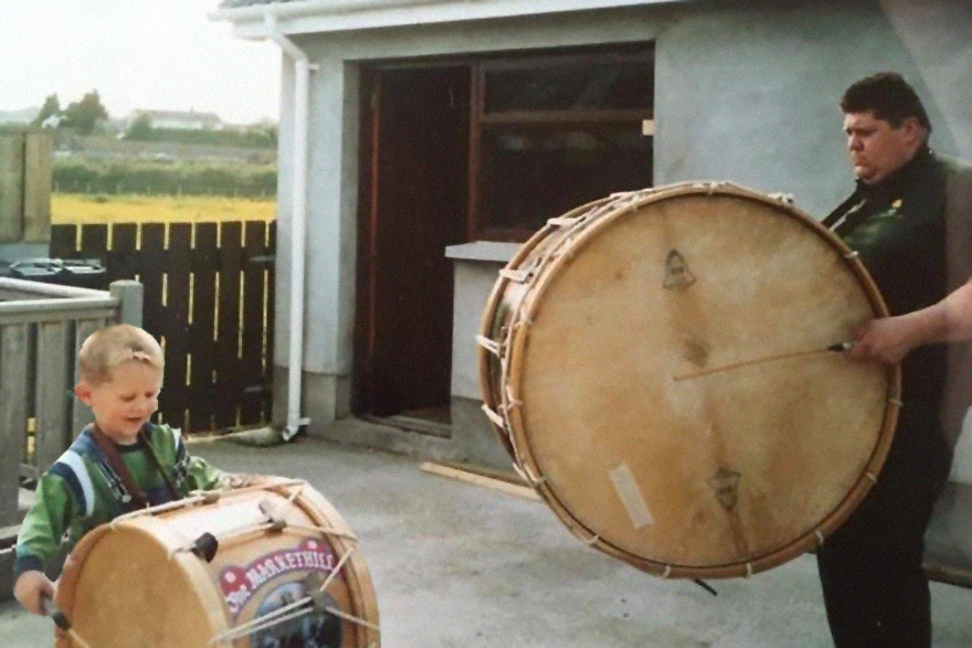 A man with a large round drum strapped to his chest watches a young boy wearing and playing a proportionally sized drum. Old color family photo.