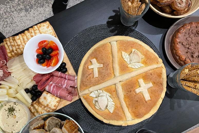 A spread of food on a dining table, including a circular loaf of bread decorated with dough shaped into roses and crosses, breadsticks, and a charcuterie board.