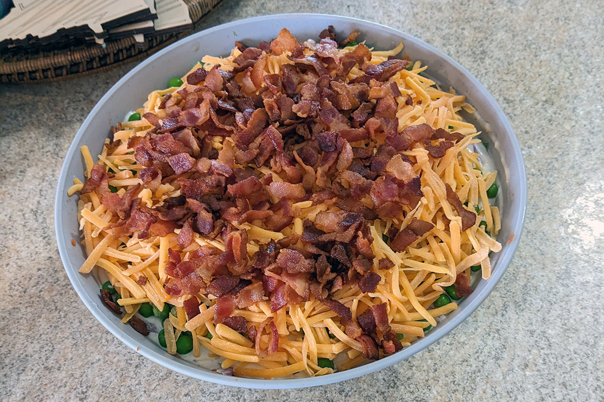 In a circular  bowl, red bacon bits and orange cheddar cheese top a salad. Green peas and white dressing peak through from below.
