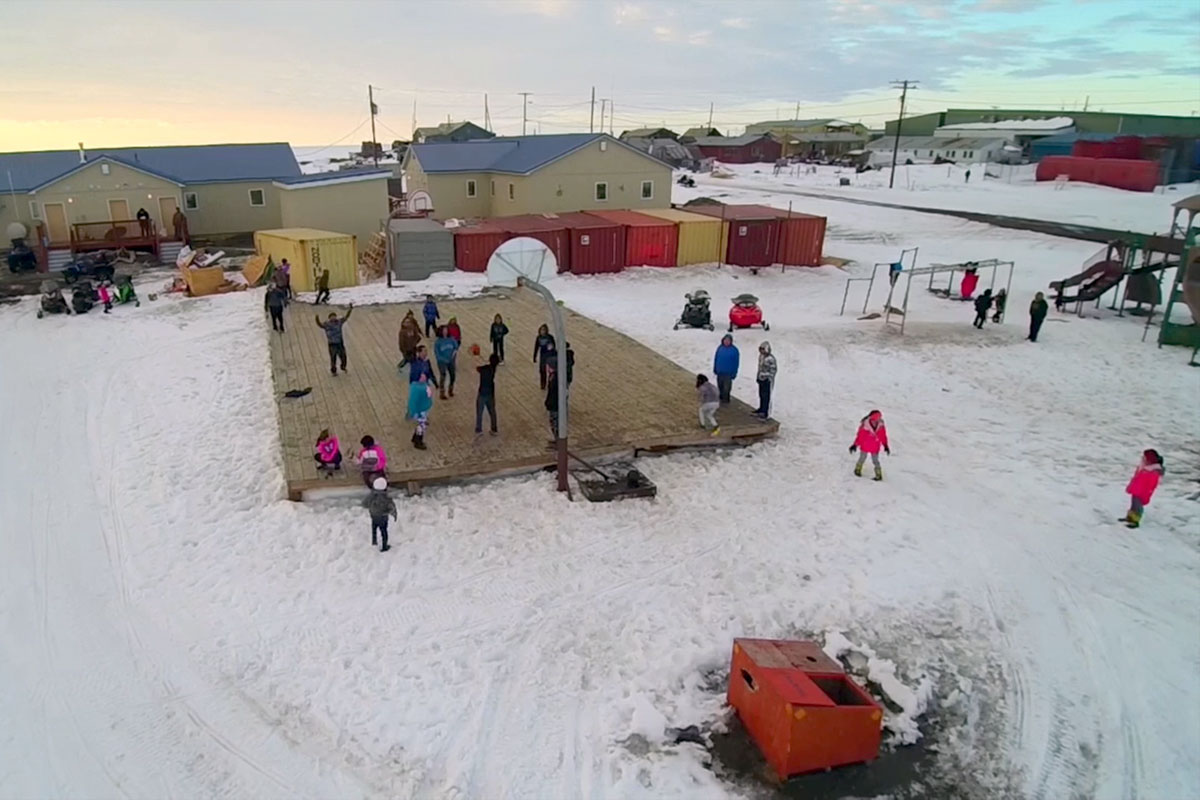 Drone shot of children and adults on and around an outdoor, wooden basketball court, raised like a stage above the snow-covered ground. In the background, storage containers, a few single-story buildings, and glow from the sun low on the horizon.