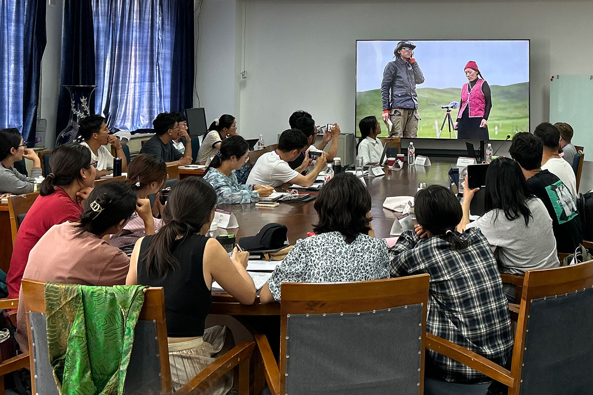 In a classroom, students sit around a large oval table, watching a video screen showing two people outdoors, a microphone set on a tripod between them. 