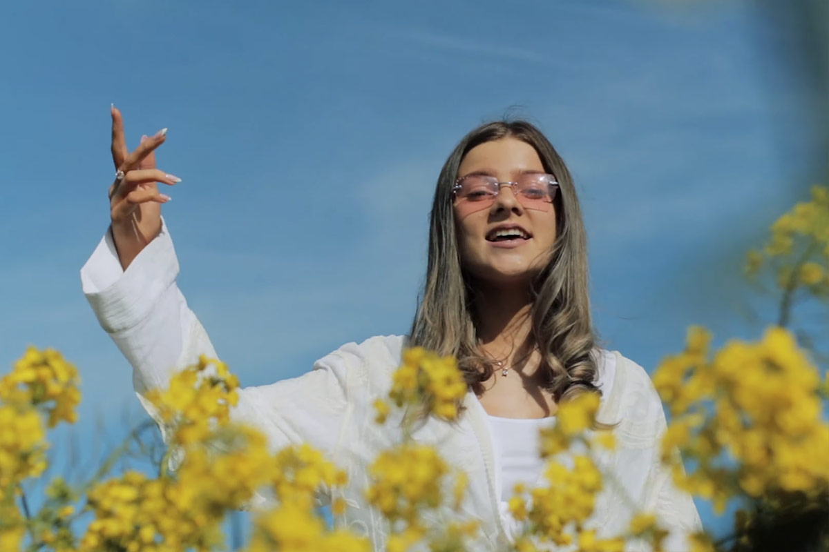 A woman in a long-sleeved white top and pink shades, with long dark hair, sings among yellow wildflowers.