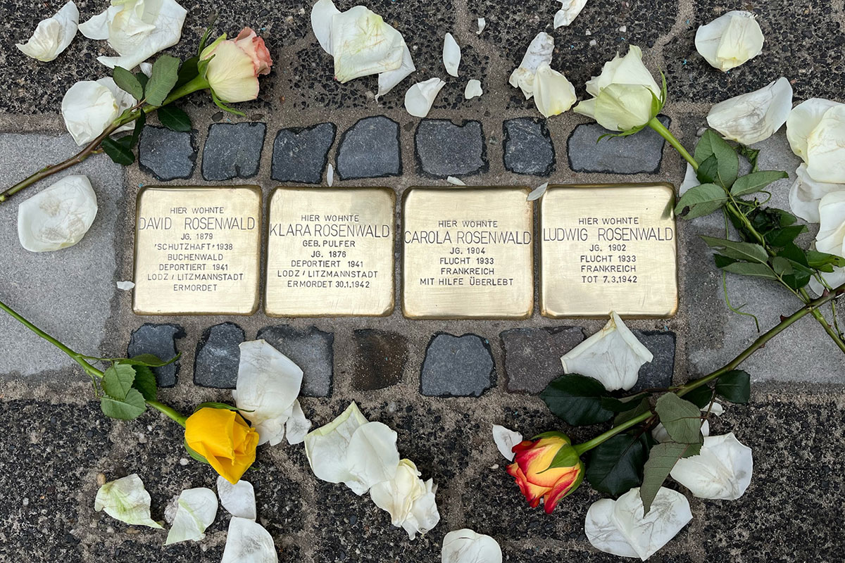 Four square brass plaques placed in a cobblestone sidewalk, frame by roses and loose white petals. Each plaque is engraved with a name and birth and death dates and other details in German. These four commemorate David Rosenwald, Klara Rosenwald, Carola Rosenwald, and Ludwig Rosenwald.