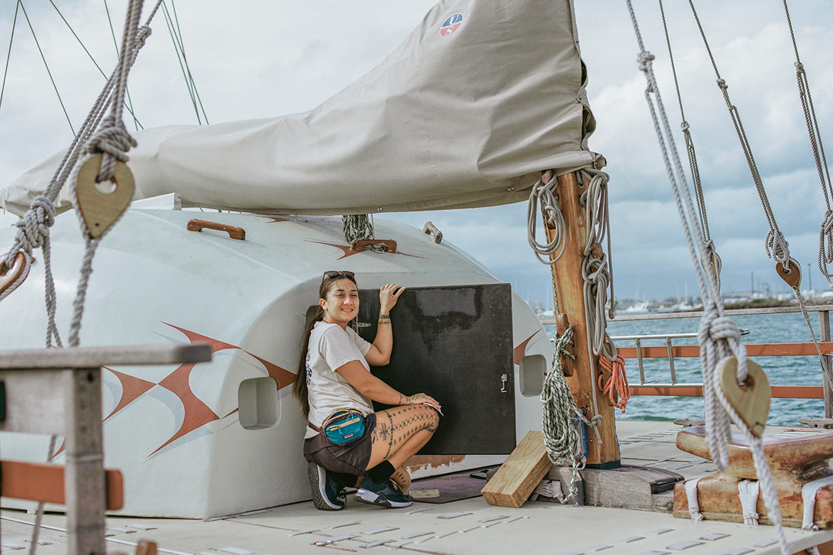 Aboard a ship, a young woman crouches on deck, smiling. She has a traditional Hawaiian pattern tattooed on her leg.