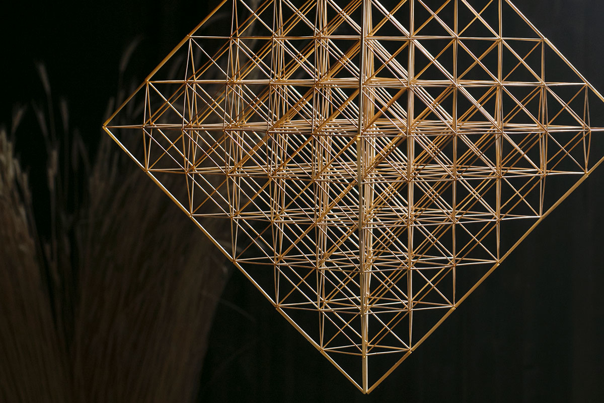 Mobile made of straw in the shape of a cube, made up of octahedrons, with a faint bouquet of straw against a black background.