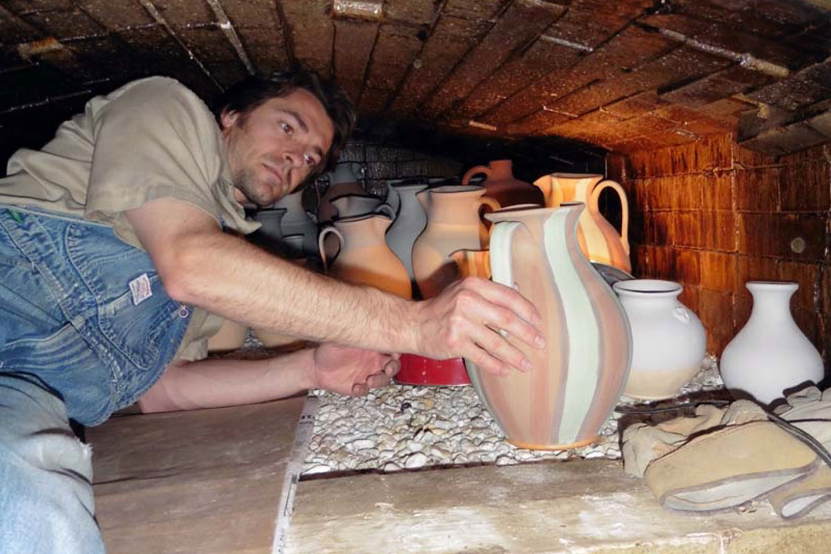 A man wearing blue overalls lies inside a brick kiln, propped up on one elbow and adjusting a gray-and-pink-striped ceramic jug. Other jugs and vases behind him await firing.