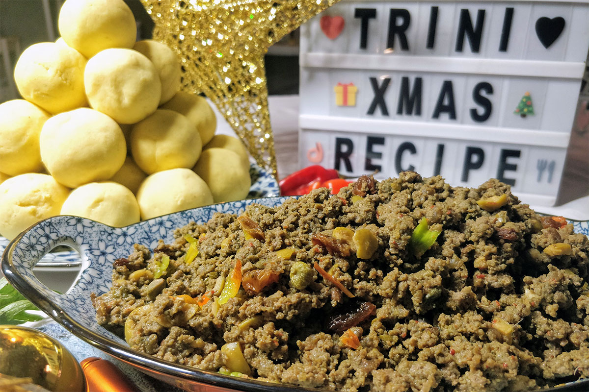 A bowl full of cooked ground meat, flecked with colorful vegetables pieces. In the background, a mound of yellow dough balls and a mini marquee-style sign that reads TRINI XMAS RECIPE.