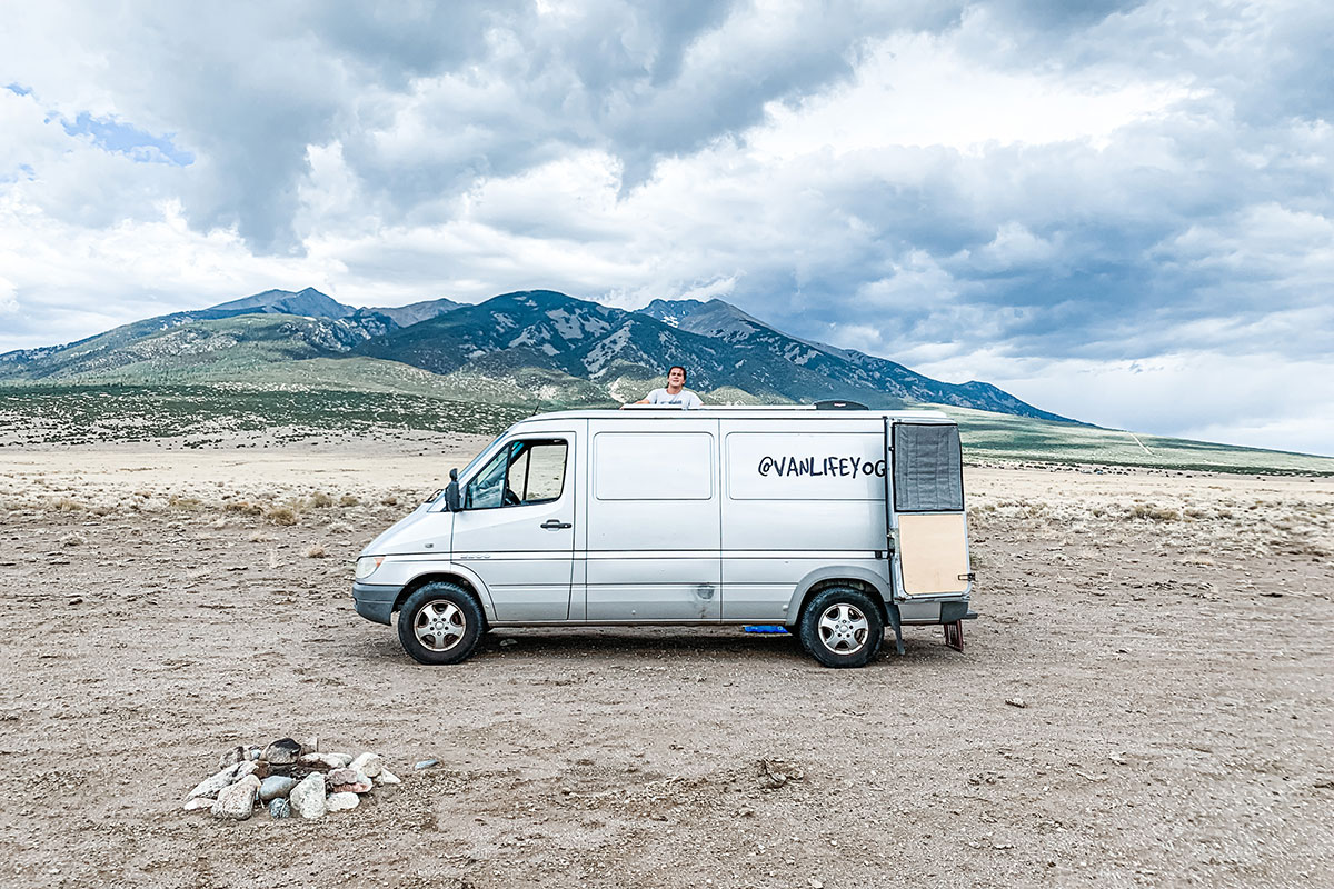A man poses with a large white van, with @vanlifeyoga spray-painted on the side. Dramatic gray clouds overhead and mountains looming in the distance.