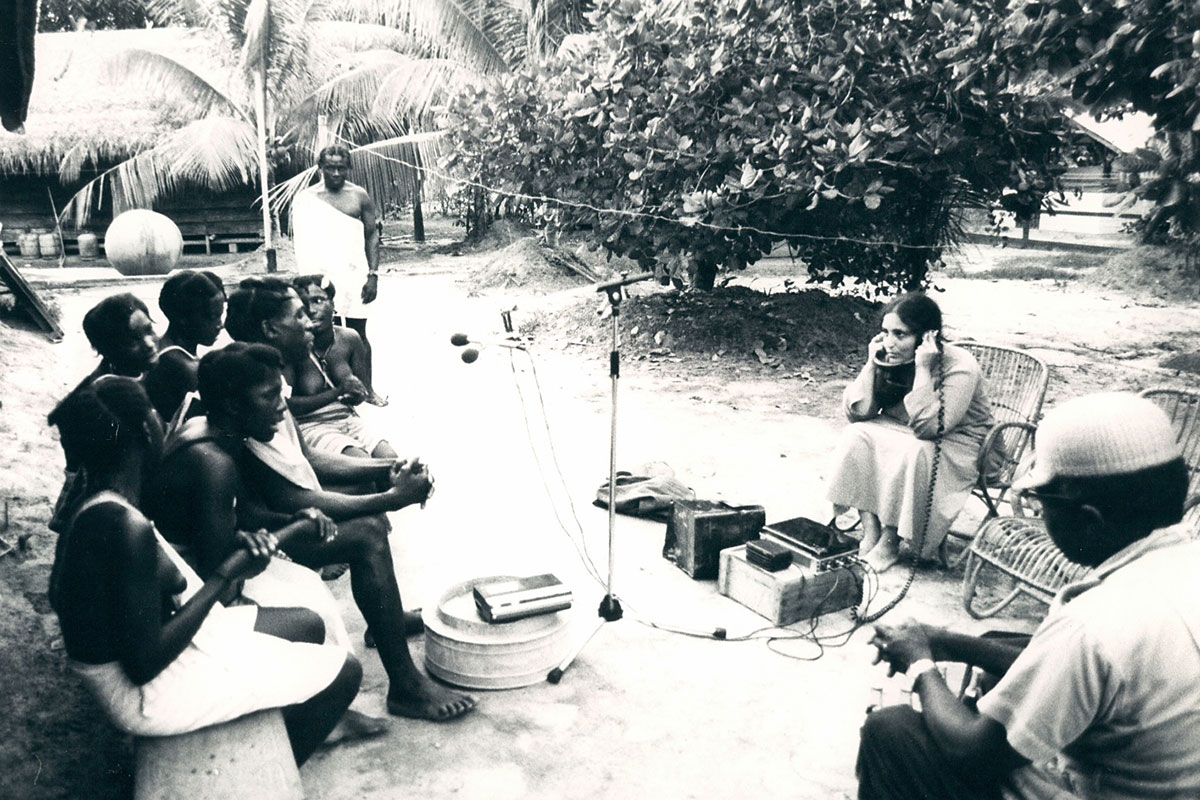 On the left, a group of Black people in white robes sing on a bench outdoors, facing a white woman recording them on the right. Between them on the ground, a microphone and recording equipment.