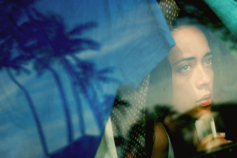 Film still of a young woman inside a vehicle, looking outside and holding a cigarette. We see reflections of palm trees in the window.