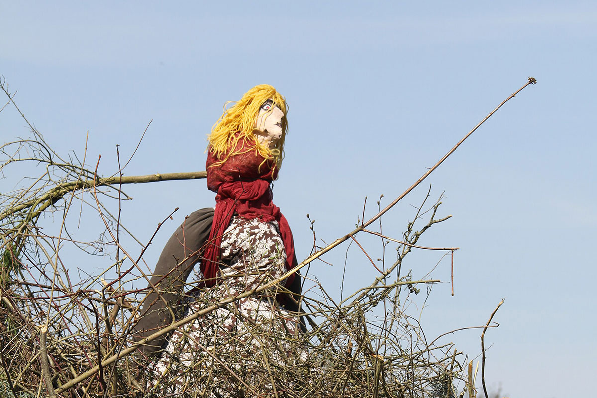 A doll of a woman with yellow hair and red dress on a pile of kindling under a blue sky.