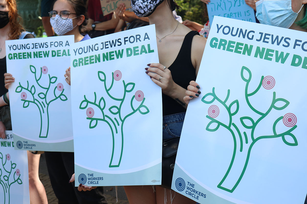 People at a rally hold up identical signs, reading Young Jews for a Green New Deal, along with an image of a tree.