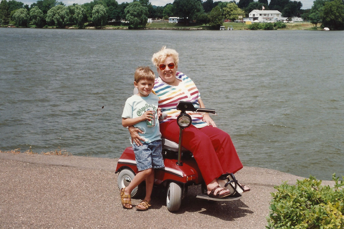 Old color photo of a young boy standing next to an older woman on an electric mobility scooter in front of a lake.