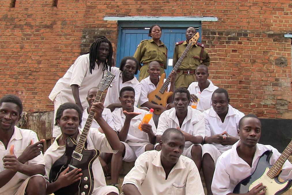Men in matching white prisoner uniforms pose, some holding up electric guitars. Two guards in green pose behind them.