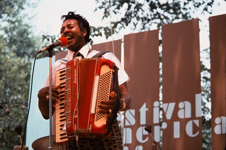 A man plays an accordion as he sings into a microphone on an outdoor stage.