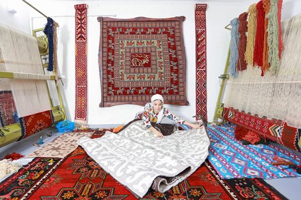 A woman sits on the ground, unrolling a woven gray and white rug. More rugs sit under her and hang behind her, and on either side are two large looms with rugs in progress.