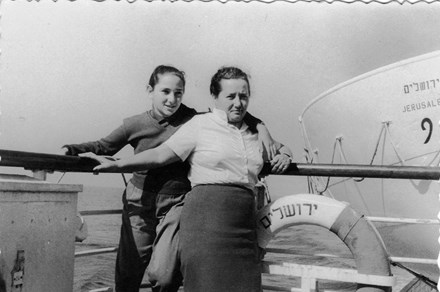 A mother and teenage daughter pose along the railing of a ship or dock. The life preserver ring next to them has text in Yiddish or Hebrew.