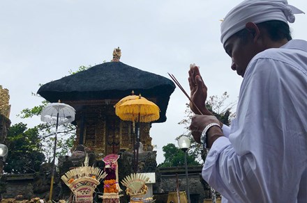 A man dressed in white shirt and head wrap holds up one hands and two sticks of maybe incense in the other. In front of him is a decorative golden temple and colorful umbrellas. 