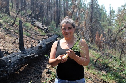 A young women holds a tuft of long grass, smiling on a forested hillside that looks like it's recovering from a fire.