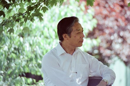 A man in white dress sits with a thoughtful expression on his face outdoors, green and red foliage out of focus behind him. Old color film photo.