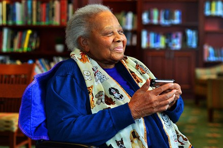 An older woman sits and smiles in a library room, holding a harmonica. Her scarf is printed with cartoon dogs.