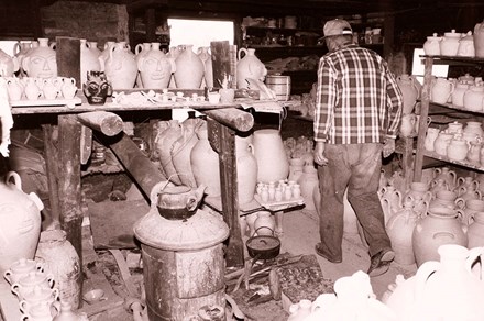 A man, facing away, walks through a workshop room. Every flat surface, including the floor, shelves, and tabletops, contains ceramic jugs in progress. Some of them have humanoid faces on the sides.