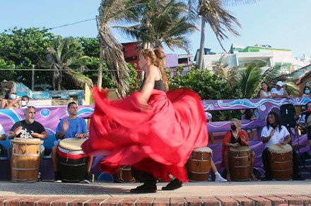 A woman in a long, flowing red skirt, blurry with motion, dances in front of a line of people seated and playing barrel-shaped drums. Palm trees sway in the background.