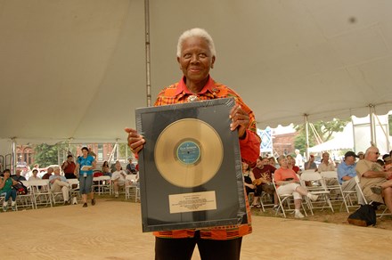 An elder woman with dark skin and short white hair holds up a framed twelve-inch gold record, smiling. Behind her is a seated crowd under a festival tent.