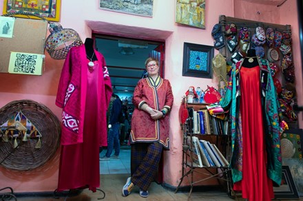A woman stands in a doorway, leaning against a pink wall in a gallery space. She looks at the camera and smiles, clasping her hands together in front of her. The room is filled with clothing pieces, hats, and art on the walls.