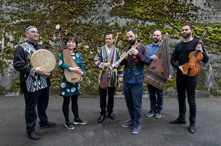 Six people pose in front of a mossy wall, holding musical instruments: a circular hand drum, a Chinese lute, long-necked bowed string instrument, saxophone, zither, and viola.
