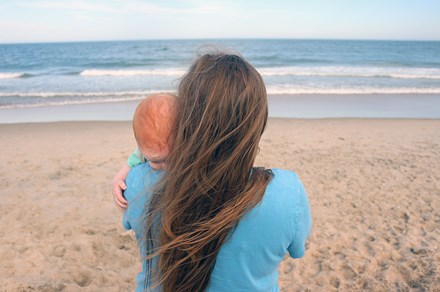 A woman standing on the beach faces the ocean, holding a small red-headed baby. Both their faces are hidden.
