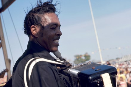 A man onstage, smiling with windswept hair and playing the accordion for a large audience. 