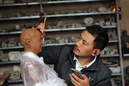 A man holds a cell phone in one hand and a wooden tool in the other, which he uses to form the face of a red clay sculpture of a person. He was wired earbuds. Behind him are tall shelves full of ceramics.