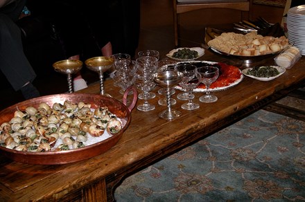 A coffee table set with a copper pan full of escargots on rock salt, wide champagne glasses, a plate of red and black caviar, and a bowl of crackers.