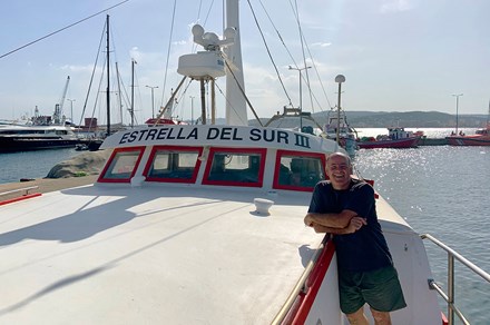 A man poses leaning against a docked white fishing boat with red trim.