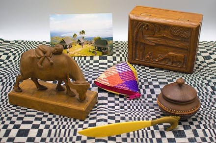 Display of objects on a black and white checkered textile: dark wooden figurine of a child riding an ox, an ivory dagger with bronze handle, a circular dark brown container, a woven coin purse in purple, red, yellow, and tan, a wooden box with an engraved scene on top, and a photograph of open-air huts and palm trees on a mountainside, a lake in the distance.