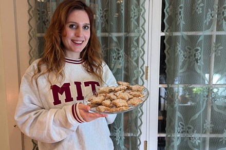 A young woman with wavy chestnut hair and at MIT sweatshirt holds up a glass plate stacked with cookies.