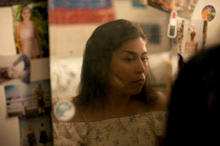 A young woman looks at herself in a bedroom mirror framed by photographs and stickers. On her cheek are two strokes of skin-toned foundation, one light and one darker than her skin tone.