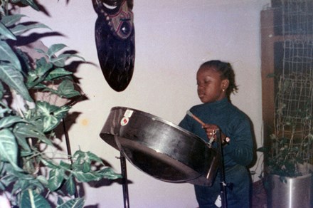 A young girl plays a steelpan drum, houseplants in the foreground and background, and a wooden mask on the wall.