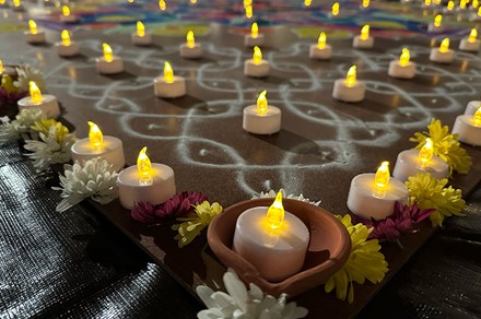 Close-up on an artwork display on the ground, of geometric patterns created with lines of white and colored sand, bordered by lit electric tea lights and white, red, and yellow flowers. The tea lights, glowing yellow, are also placed throughout the sand patterns.