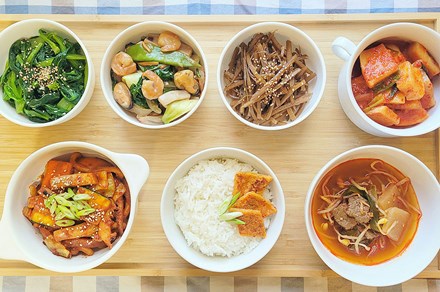 Seven bowls of various side dishes: wilted greens with sesame seeds, shrimp and vegetables, rice, soup with beef and bean sprouts, and more.